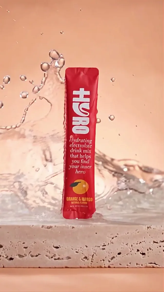 A red HYRO Orange & Mango pack surrounded by an arc of water on a warm peach background.